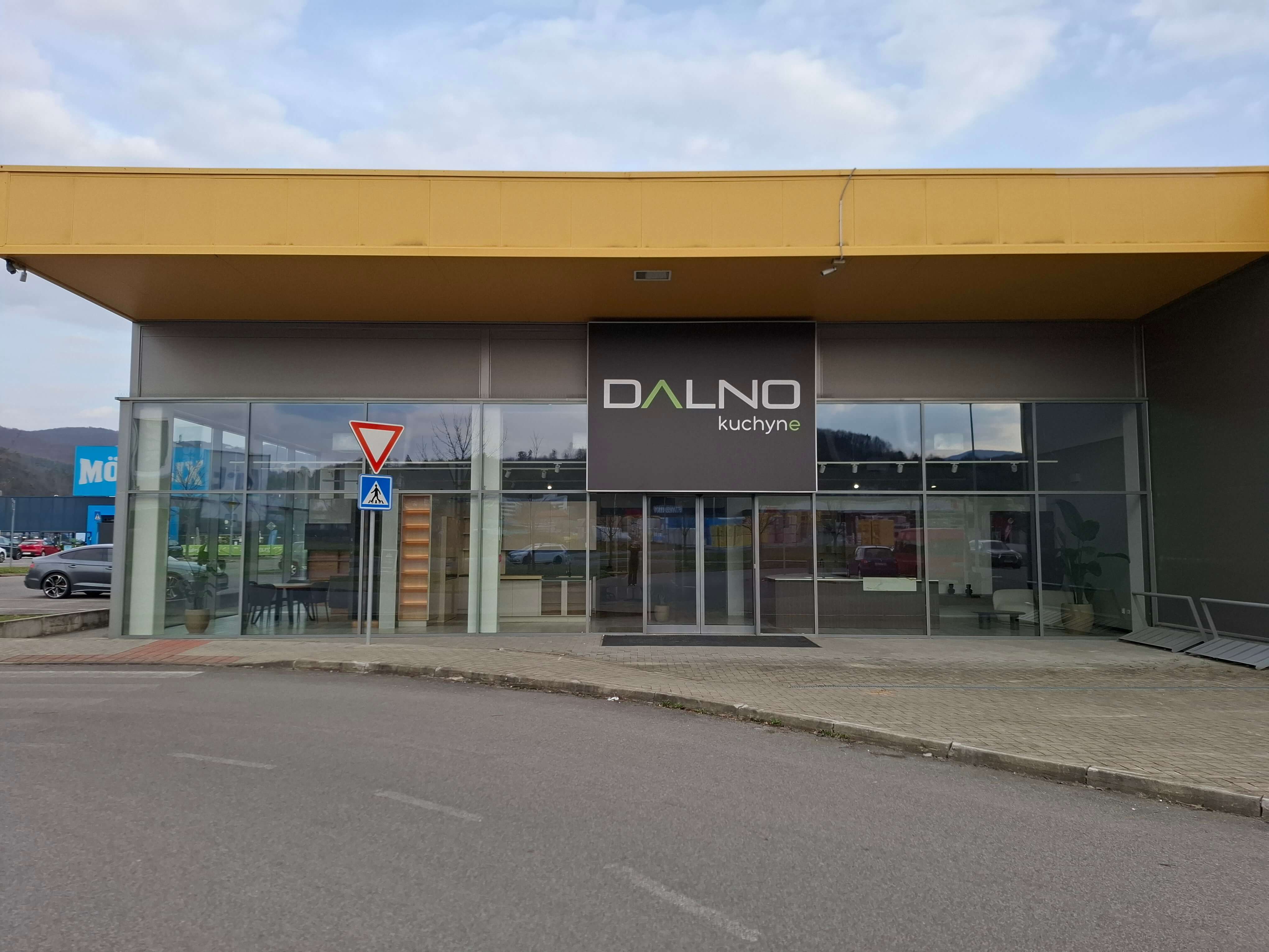 Exteriér showroomu Dalno Banská Bystrica s moderným dizajnom budovy, presklenými stenami a logom značky na fasáde. Pred vstupom je upravený priestor a viditeľné parkovacie miesta.